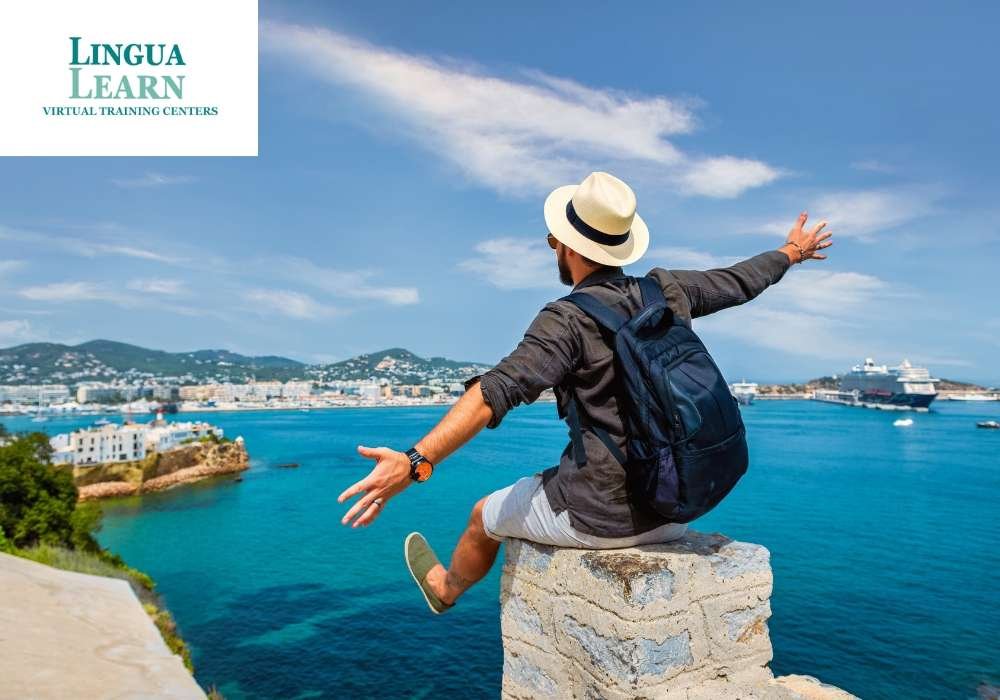Person in a hat sitting on a wall, arms outstretched, overlooking a blue sea