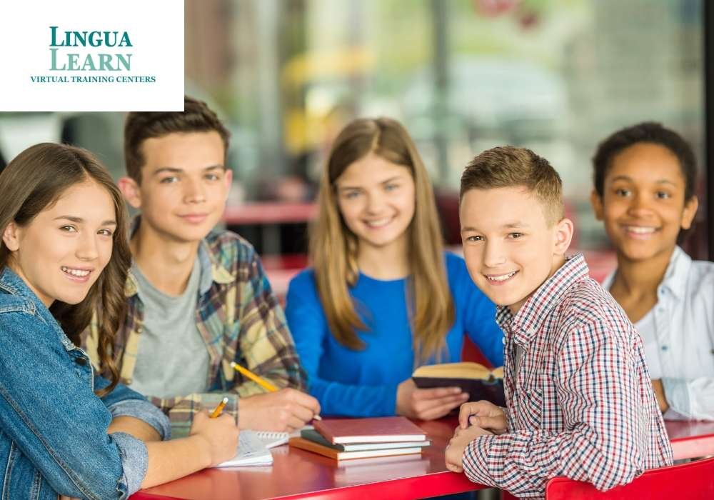 Five smiling students sitting at a table with notebooks and pens